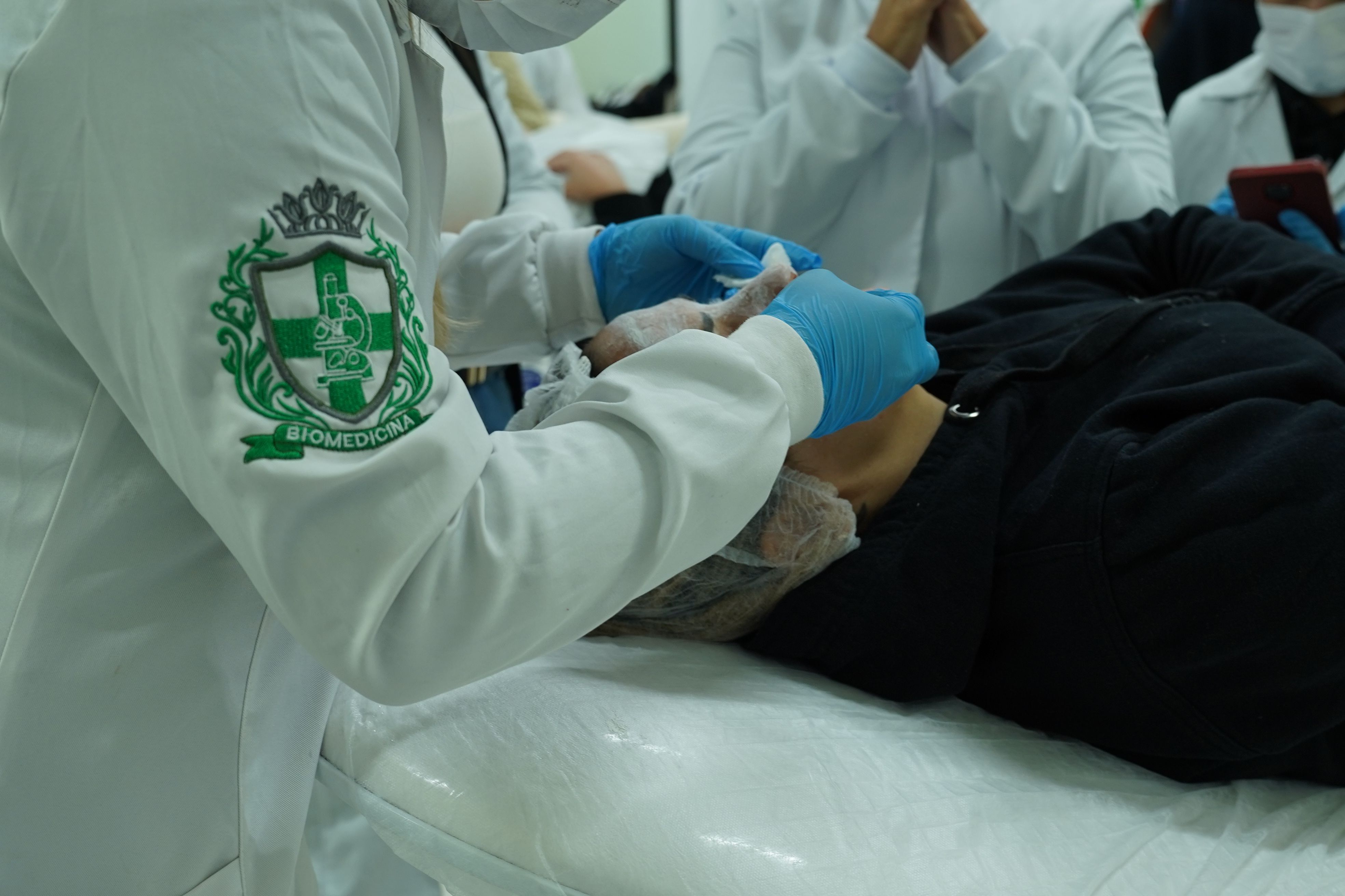 Estudante de jaleco aplicando procedimento em paciente deitado, durante aula prática na área da saúde, com outros alunos ao fundo.