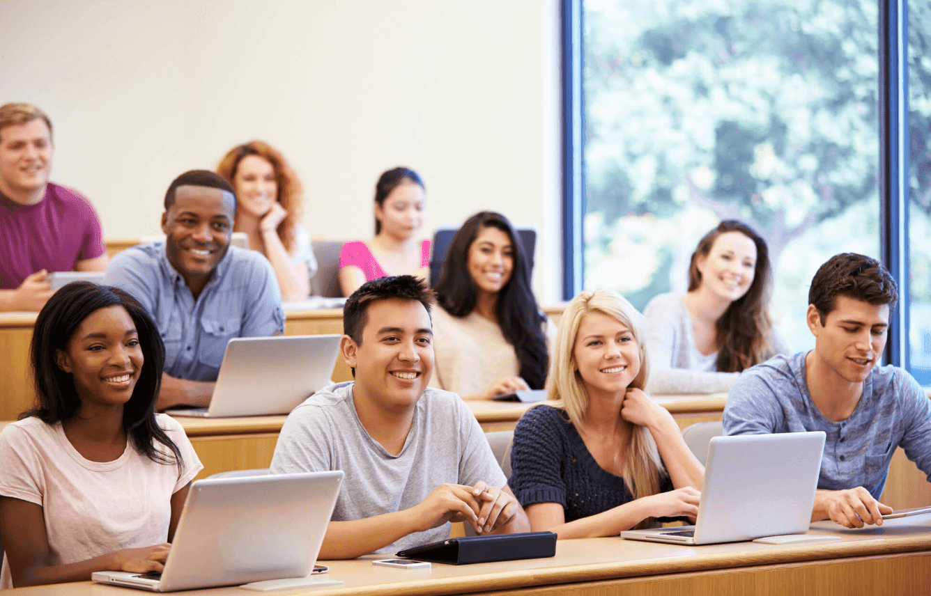 Alunos sorrindo em sala de aula com notebooks, sentados em carteiras dispostas em degraus, em ambiente iluminado e moderno.