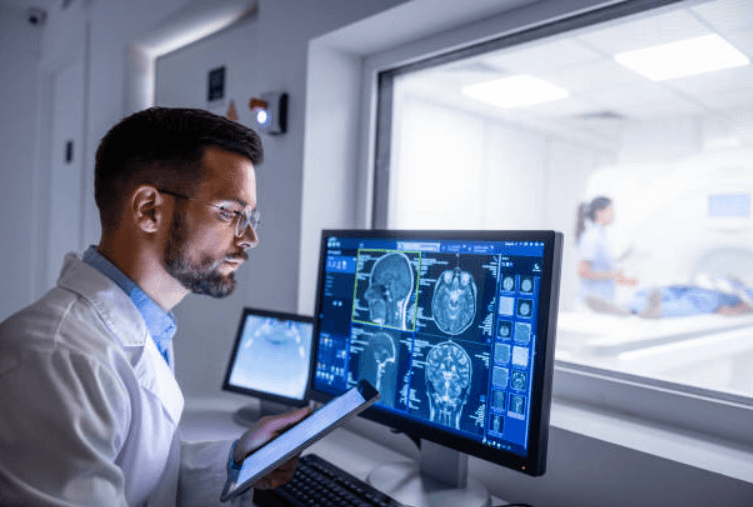 Profissional de saúde analisando exames de ressonância magnética em um computador, enquanto paciente realiza exame em máquina de imagem ao fundo.